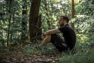 Side view of thoughtful man sitting against trees at forest