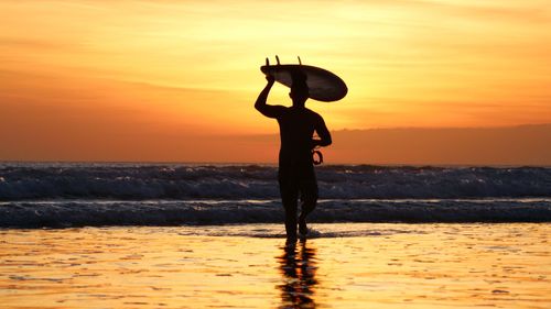 Silhouette mid adult man with surfboard on head walking in sea against sky during sunset
