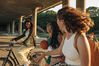 Teenage girls looking at smiling female friend riding bicycle under bridge