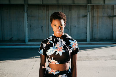 High angle of charismatic confident african american female athlete in flowered sportswear looking at camera with challenge while standing alone on street in sunbeams against concrete wall in city