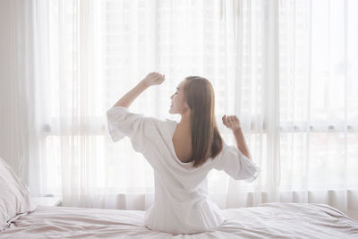 Rear view of young woman with arms raised sitting on bed against window at home