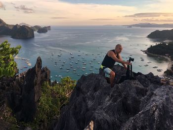 Man sitting on rock at beach against sky during sunset