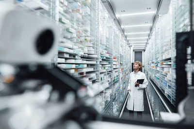 Female pharmacist with digital tablet searching medicine seen through machinery at pharmacy store