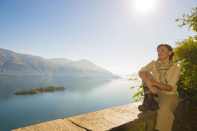 Young woman sitting on retaining wall by alpine lake against sky