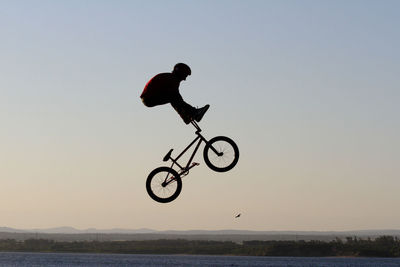 Man with bicycle jumping over sea against clear sky