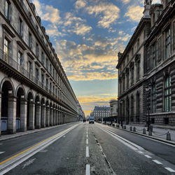 Road amidst buildings in city against sky at sunset