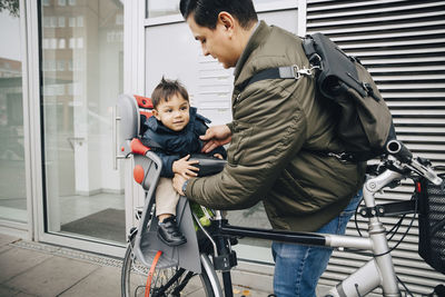 Father holding son sitting in safety seat of bicycle on sidewalk in city