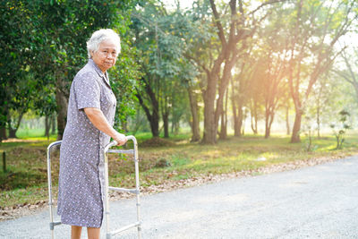 Portrait of disabled woman standing with mobility walker on road against trees
