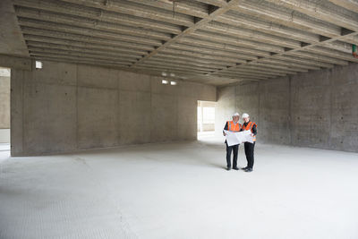 Two men with plan wearing safety vests talking in building under construction