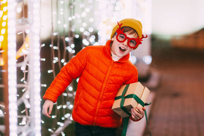 A cheerful boy in an orange jacket and funny glasses walks through the evening city with festive 