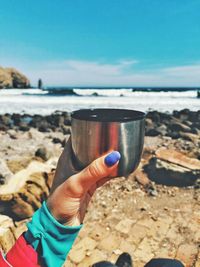 Close-up of hand holding drink at beach against sky