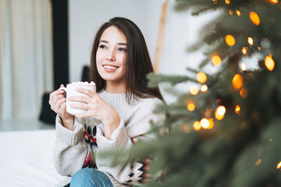 Young asian woman  with cup of cocoa with marshmallow in room with christmas tree at home