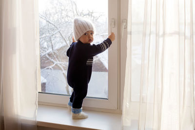 Child in a warm blue jumpsuit is on window in winter white knit hat