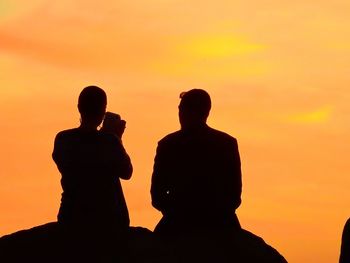 Silhouette people sitting on rock against orange sky