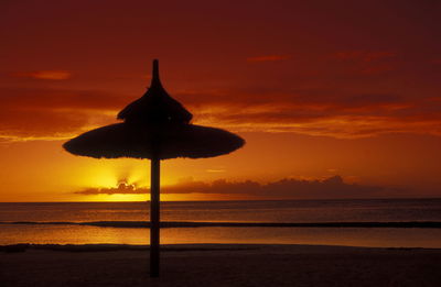 Silhouette thatched roof on beach against orange sky