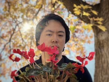 Close-up of young asian man with red flowering cyclamen plant against autumn foliage and tree.