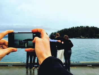 Cropped image of hands photographing friends by lake against sky