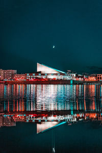 Illuminated buildings by river against sky at night