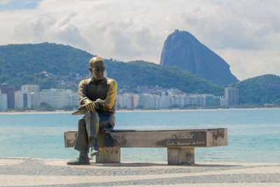 Rear view of man sitting by sea against mountains