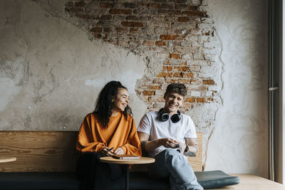 Happy male and female entrepreneurs laughing while sitting on bench against brick wall in office