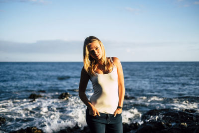 Portrait of young woman standing at beach against blue sky during sunny day