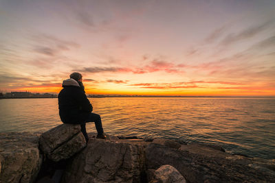 Man sitting on rock by sea against sky during sunset