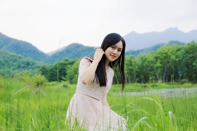 Portrait of smiling beautiful woman kneeling grassy field against mountains