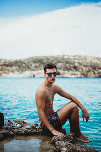 Side view portrait of shirtless man wearing sunglasses sitting on rock at beach