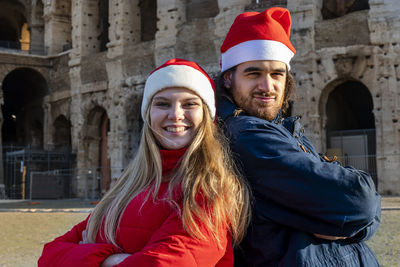 A lovely young couple taking a picture in front of the colosseum with red santa hats. 