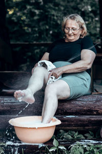 Elderly woman in glasses washing her feet in basin of foam in backyard on hot summer day