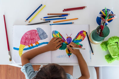 High view of a little girl sitting on the table and painting pictures of animals.