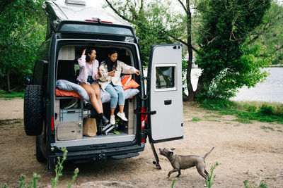 Traveling young multiracial girlfriends drinking beer and giving command to dog while having fun and resting together in camper van parked near river in countryside