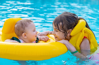 Smiling girl in life jacket looking at sister with inflatable ring in swimming pool