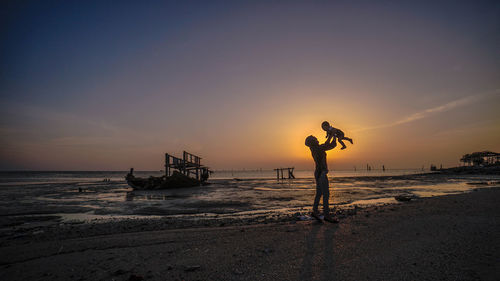 Woman holding baby while standing at beach against sky during sunset