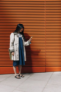 Woman using smart phone while standing in front of orange wall