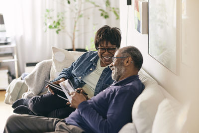 Happy senior woman with digital tablet talking to man sitting on sofa at home