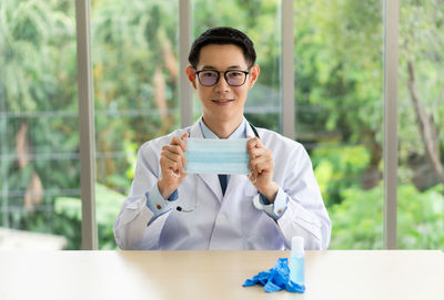 Portrait of doctor holding flu mask sitting against plants