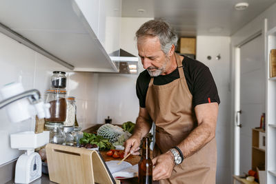 Mature man reading book while standing in kitchen at home