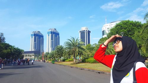 Young woman shielding eyes while wearing hijab on road in city against sky