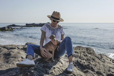 Woman sitting with dog on rock at sea shore against sky