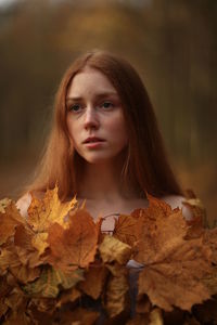 Thoughtful young woman covered with leaves at forest during autumn