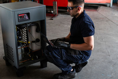 Adult man in goggles using laptop to program broken machine during work in modern workshop