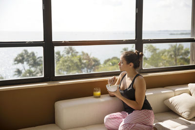 Woman looking through window while eating breakfast on sofa at home