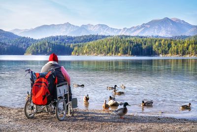 Rear view of person sitting on wheelchair at lake against mountains