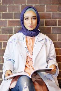 Portrait of beautiful young woman sitting with book against brick wall