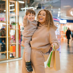 Smiling mother carries her little happy son in her arms, holds shopping bags in her hands and walks