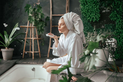 Beautiful young woman enjoys her bathroom and blowing bubbles soap wearing bathrobe and towel
