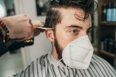 Male hairdresser haircutting hair to a young bearded man wearing a face mask due to coronavirus 