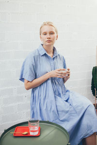 Portrait of young woman with coffee sitting against wall