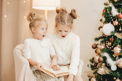 2 blonde girls are sitting on an armchair by the christmas tree and reading a book. 